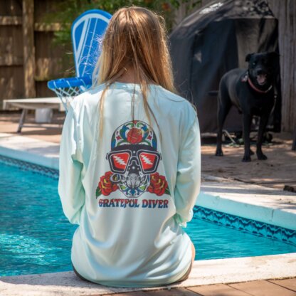 A woman sits on the edge of a pool wearing a Grateful Diver Sugar Skull UV Shirt, looking at a dog.