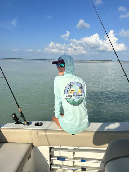 A woman sitting on a boat, wearing the Salty Soul Charters UPF 50+ Hoodie and holding a fishing rod.